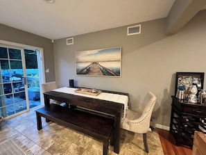 A dining area with a dark wooden table, a white chair, and a bench. There is a painting on the wall and a small shelf with various items. - Summer getaway (East Quogue)