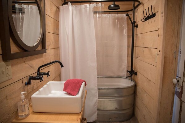 Un baño pequeño y rústico con paredes de madera, un lavamanos blanco con una toalla roja, una ducha y un espejo.