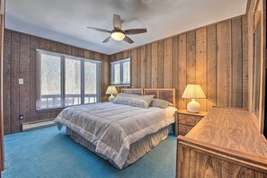A bedroom with a wooden paneled wall, a bed with a headboard, two windows with blinds, a ceiling fan, and a nightstand with a lamp.
