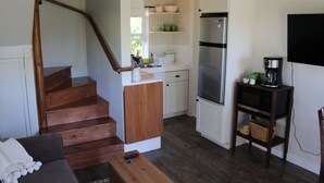 Eine kompakte Küche mit integriertem Kühlschrank, einer Mikrowelle und einer Kaffeemaschine auf einem Holztisch. Eine Treppe führt zu einem Wohnbereich mit einer Couch und einem Couchtisch.
