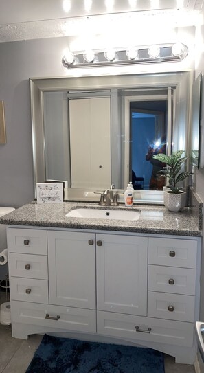 Features a spacious vanity with a large mirror, sink, and countertop, plus a cabinet and drawers for ample storage.