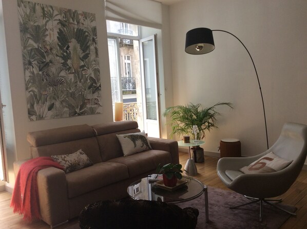 A living room with a sofa, armchair, coffee table, and a floor lamp. There is a large window with a view of buildings outside.