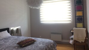 Ett sovrum med en säng, en hängande lampa, ett fönster med persienner, ett element och en vägg med inramade konstverk.