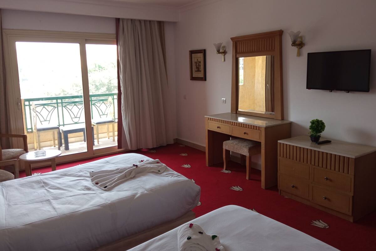A hotel room with two beds, a wooden dresser, a vanity with a mirror, a television, and a balcony with a chair and table.