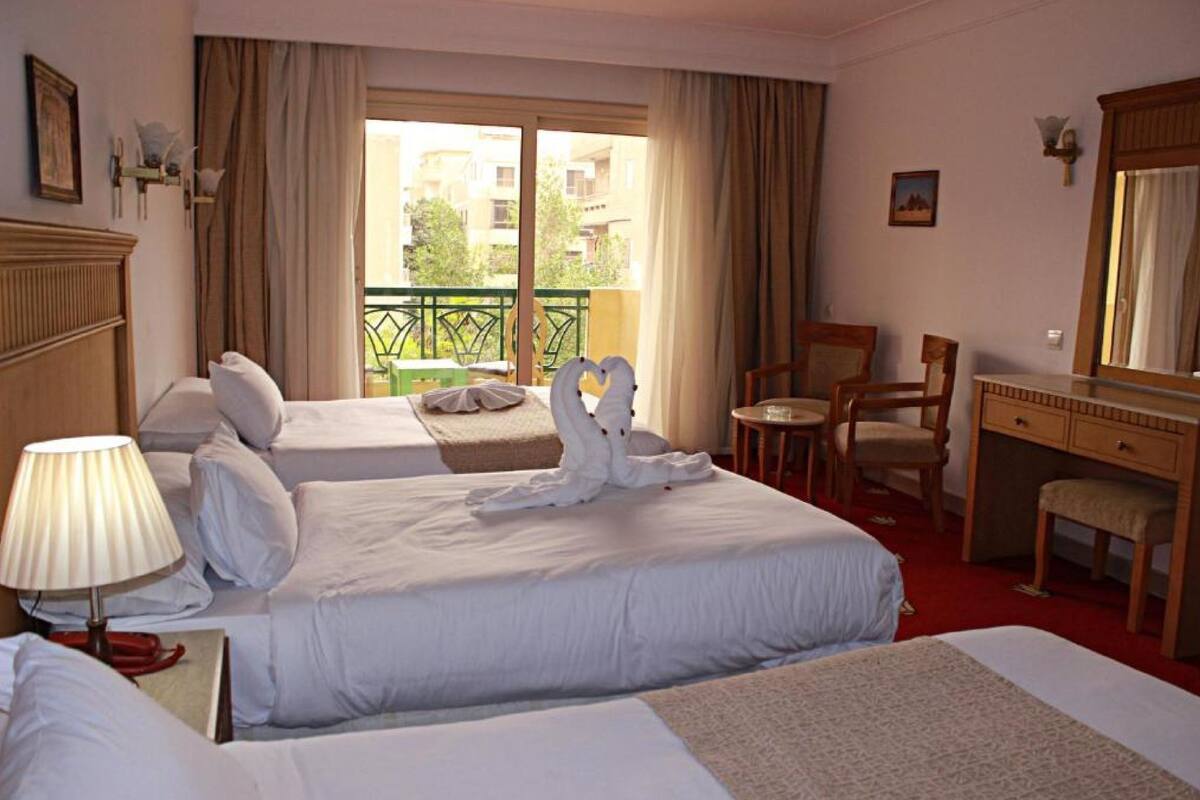 A hotel room with two beds, a desk, a chair, and a balcony with a view of buildings.