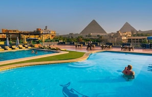 A swimming pool with a dolphin design, surrounded by lounge chairs and umbrellas, with the pyramids in the background.