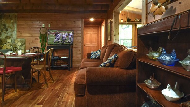 A cozy room with wooden walls, a brown sofa, a dining table with chairs, and a TV on the wall. - The Tulip Poplar Cabin - Perfect for Two People (Topton)