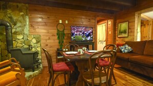 A cozy cabin interior with a stone fireplace, wooden walls, a dining table with chairs, a television, and a couch. - The Tulip Poplar Cabin - Perfect for Two People (Topton)