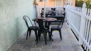 A patio with a round table and four chairs, a white picket fence, and a building in the background. - Rigo’s Apartment  (South Gate)