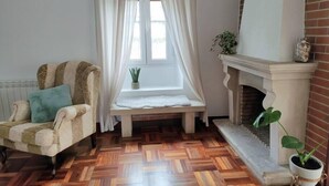 A cozy room with a wooden herringbone floor, a cushioned armchair with a teal pillow, a bench by a window with sheer curtains, and a fireplace with a potted plant. - Apartamento Lurra Barrika (Barrika)