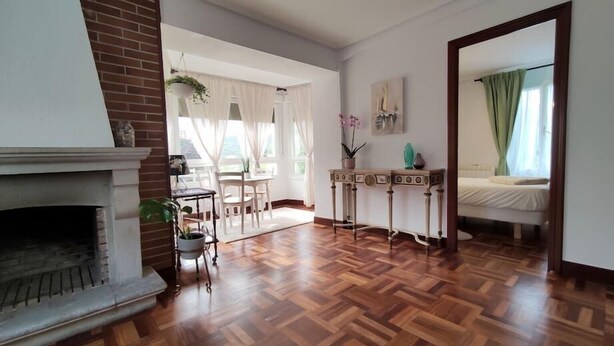 A spacious living room with a fireplace, wooden parquet flooring, a dining area with a table and chairs, and a bedroom with a bed and curtains. - Apartamento Lurra Barrika (Barrika)