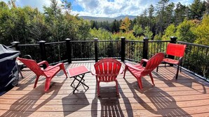 Eine Holzterrasse mit roten Adirondack-Stühlen und einem kleinen Tisch, mit Blick auf eine bewaldete Landschaft mit einem Berg in der Ferne.