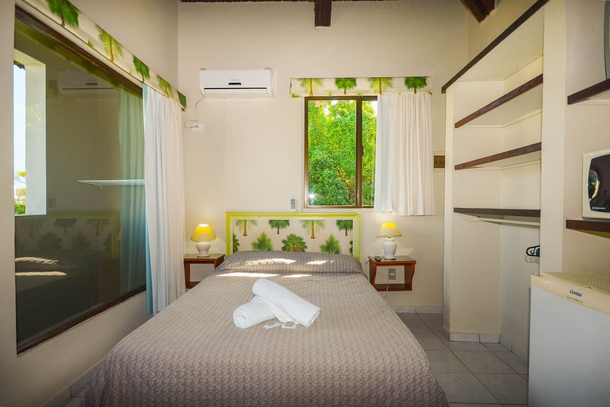 A bedroom with a bed, a window with curtains, a wall-mounted air conditioner, and a small table with a lamp.