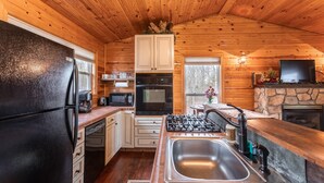 A wooden cabin kitchen with a refrigerator, stove, sink, cabinets, and a stone fireplace.