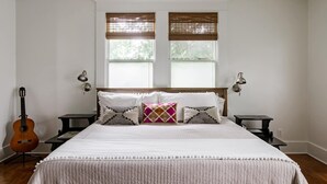 A bedroom with a bed, two nightstands, a guitar, and two windows with blinds. - Elegant Historic Estate: Professionally Designed (Austin)