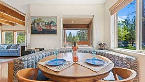 A dining area with a round wooden table, two blue plates, and a bottle of wine. There is a painting on the wall and a window with a view of trees. - Mountain Breeze - Pet Friendly Read Our Reviews (Weed)