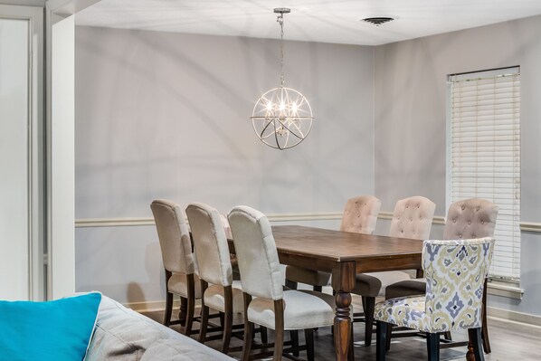 A dining area with a wooden table and upholstered chairs, a hanging light fixture, and a window with blinds. - Cheerful, Relaxing Home Away From Home! (Jackson)