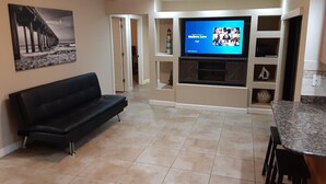 A modern living room with a black leather sofa, a flat-screen TV, a kitchen area with a granite countertop, and a hallway leading to another room.