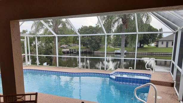 A pool with a view of a lake and trees, surrounded by a covered area with chairs and a table.