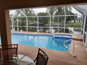 Ein Pool mit Blick auf einen See und Bäume, umgeben von einer überdachten Fläche mit Stühlen und einem Tisch.