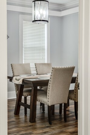 A dining area with a wooden table and four upholstered chairs, a chandelier, and a window with blinds. - Royal Chateau in the heart of Fondren! (Jackson)