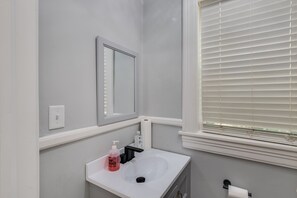 A bathroom with a white sink, a mirror, a window with blinds, and a toilet paper holder. - Royal Chateau in the heart of Fondren! (Jackson)