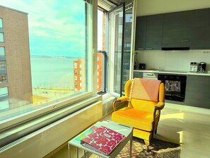 A modern living room with a large window, a yellow armchair, a small table with a magazine, and a kitchen area with cabinets and appliances. - The Cutest Studio in Central Tampere (Tampere)