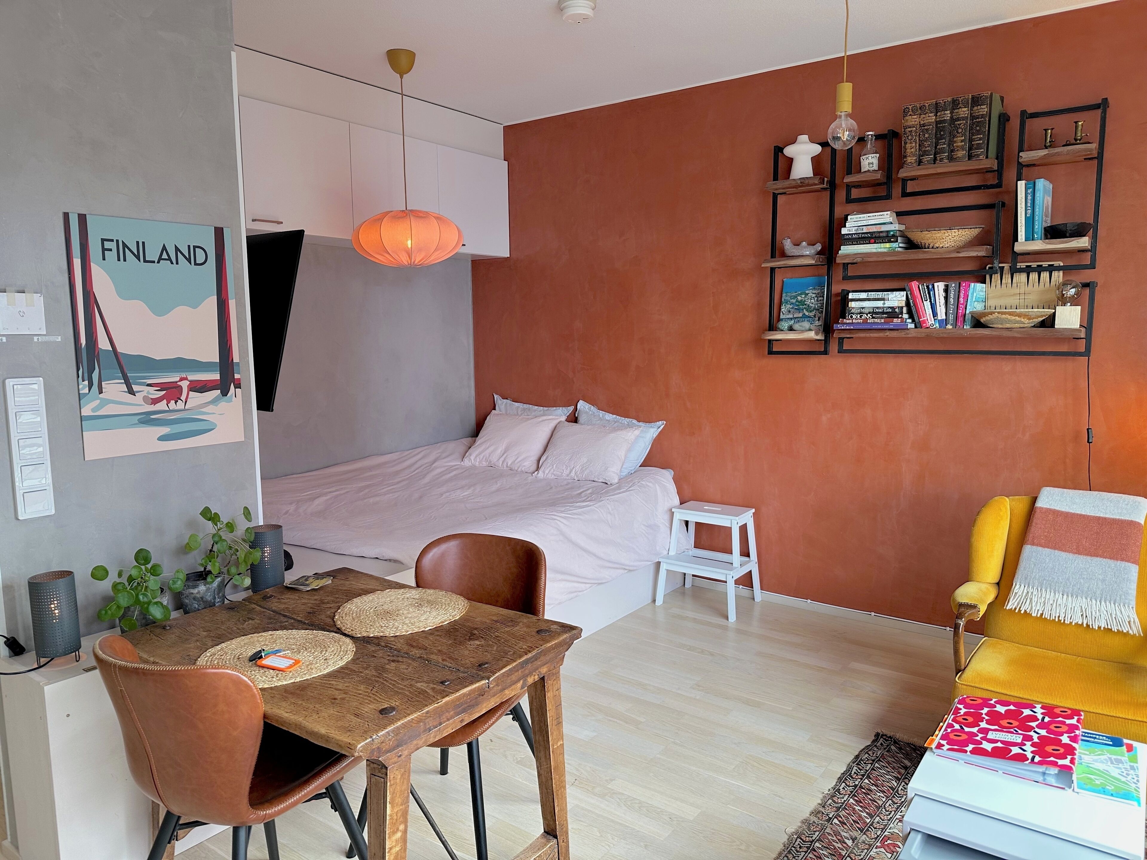 A modern bedroom with a bed, a small white table, and a yellow armchair. There is a bookshelf with books and decorative items.