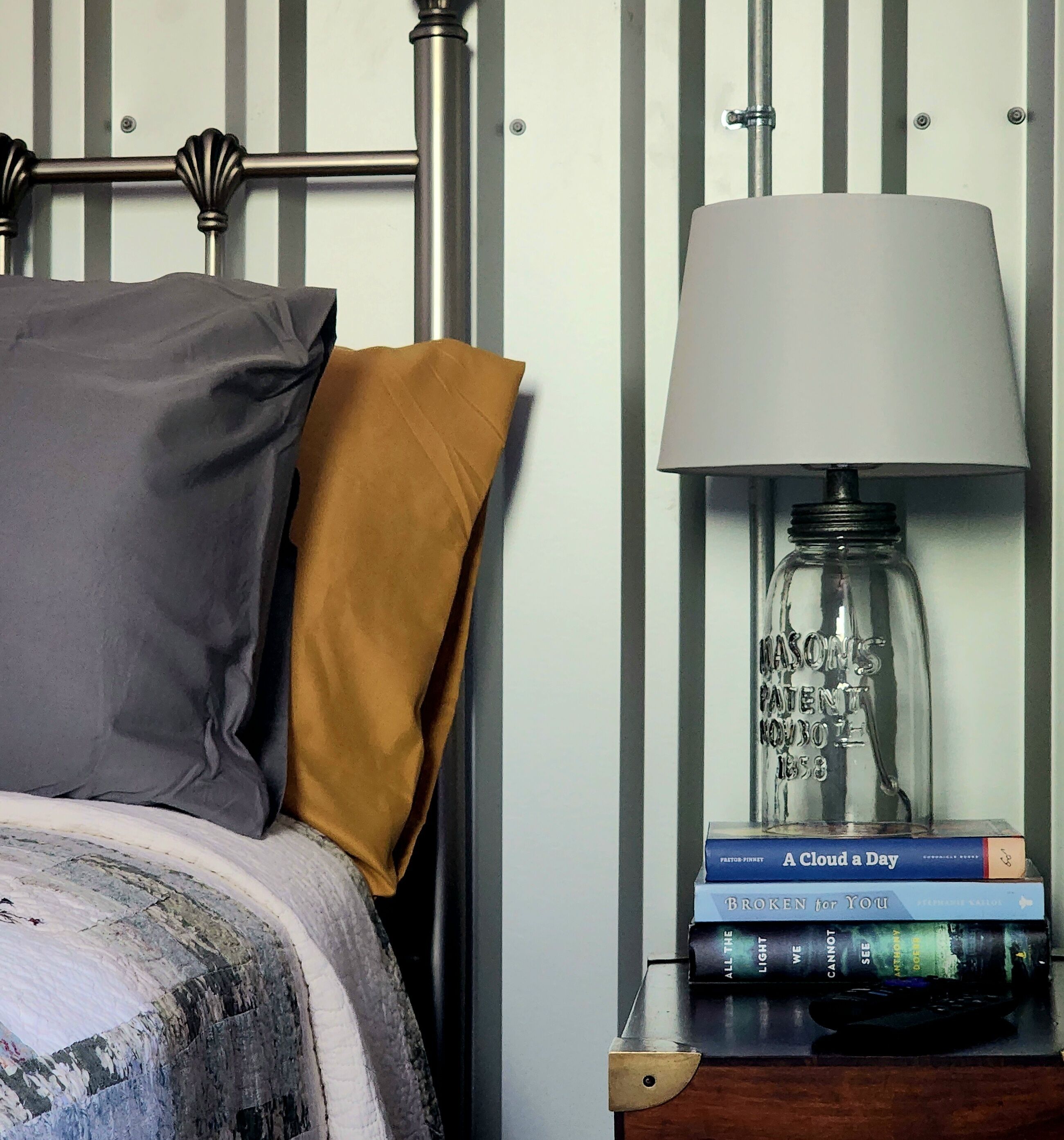 A bed with a grey pillow and a mustard-colored pillow, a bedside table with a lamp, and a stack of books.