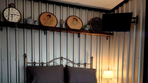 Una cama con estructura metálica y colcha, un reloj de pared y una estantería con objetos decorativos.