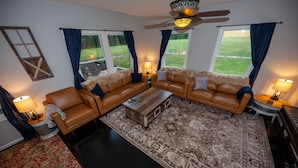 Un salon comprenant un canapé d’angle, une table basse et un tapis à motifs. Il y a deux fenêtres avec des rideaux, un ventilateur de plafond et une lampe sur une table d’appoint.