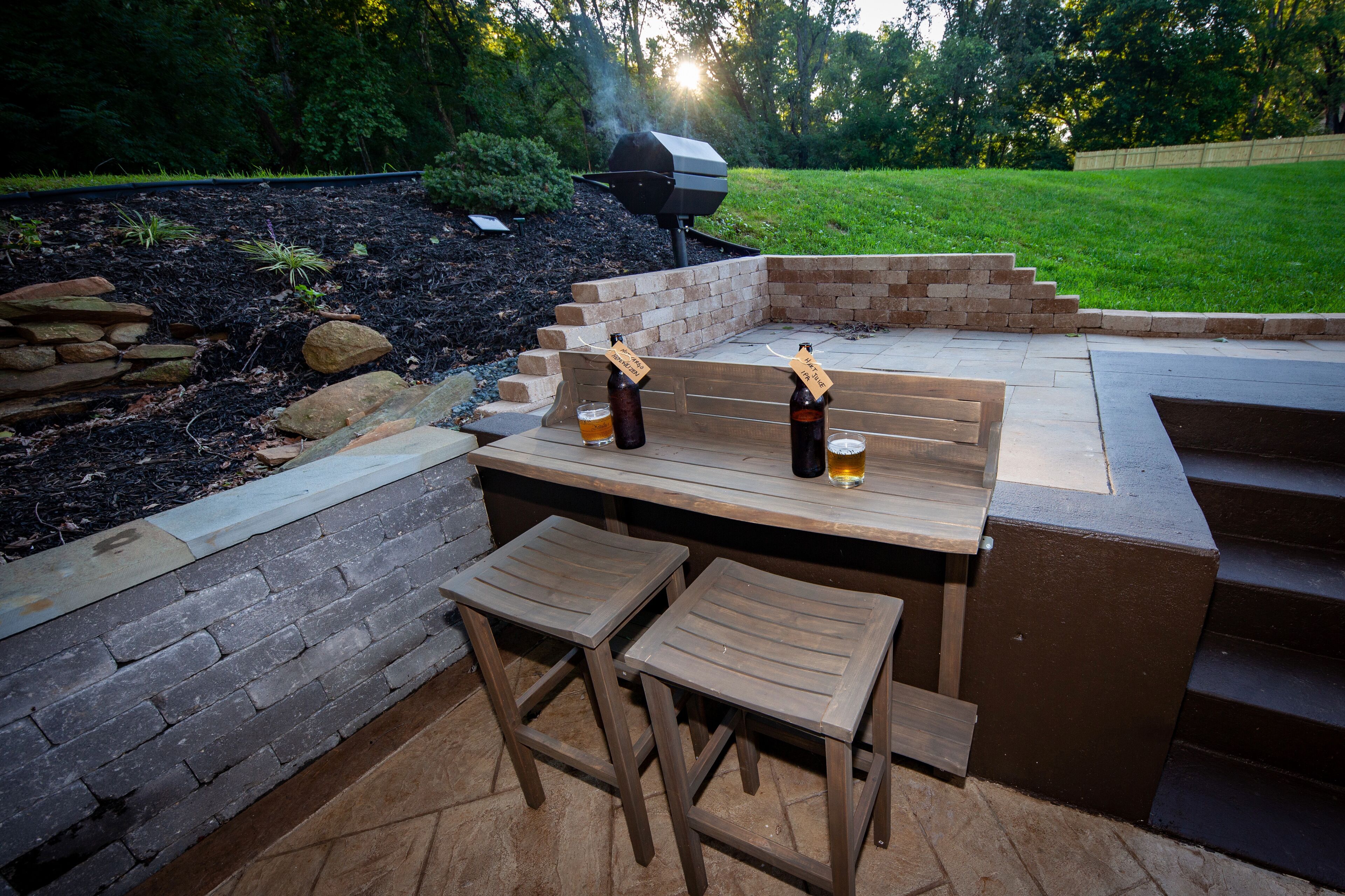 An outdoor patio with a wooden table and two stools, a barbecue grill, and a stone wall.