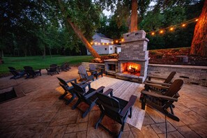 Un patio al aire libre con una chimenea de piedra, sillas de madera y luces colgantes.