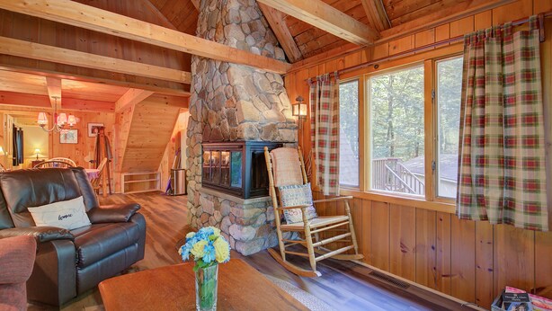 A cozy cabin interior with a stone fireplace, wooden beams, a rocking chair, and a large window with plaid curtains.