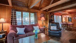 A cozy cabin interior with a wooden ceiling, a red sofa, a leather armchair, a dining table, and a clock on the wall.