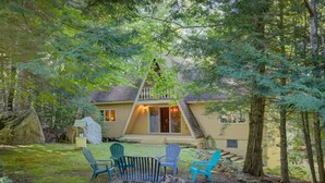 A cabin with a porch, surrounded by trees. There are four Adirondack chairs on the porch.