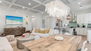 A spacious living room with a wooden dining table, white sofas, and a large painting of a beach. - Resort Pool · 4BR/3BA · 30A · Golf Cart + 8 Bikes (Santa Rosa Beach)