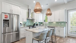 A modern kitchen with white cabinets, stainless steel appliances, a central island with bar stools, and woven pendant lights. - Resort Pool · 4BR/3BA · 30A · Golf Cart + 8 Bikes (Santa Rosa Beach)