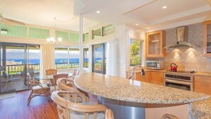 A kitchen with a large island, bar stools, a view of the ocean, and a dining area with chairs. - Kapalua Ridge 2723-24- Full/Island Sunset Views (Lahaina)