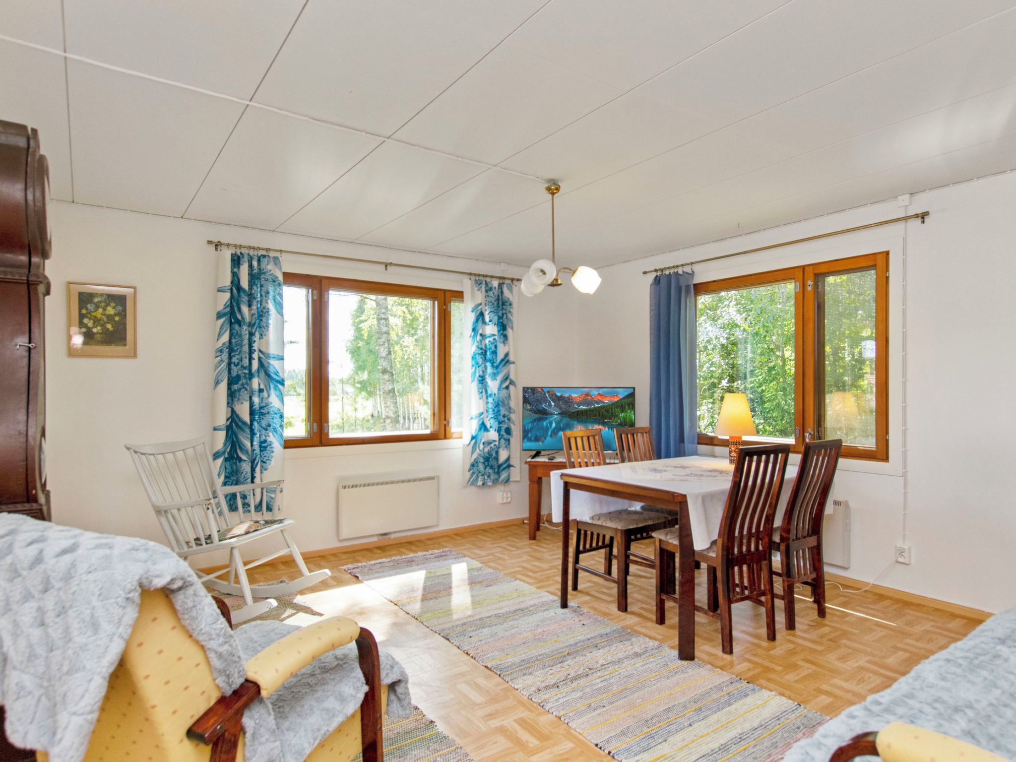 Une pièce comprenant une table à manger, des chaises, un fauteuil à bascule, un radiateur et de grandes fenêtres avec des rideaux.