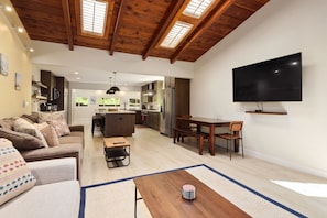 Ampla sala de estar com teto de madeira, uma grande TV de tela plana, uma mesa de jantar e cozinha ao fundo.