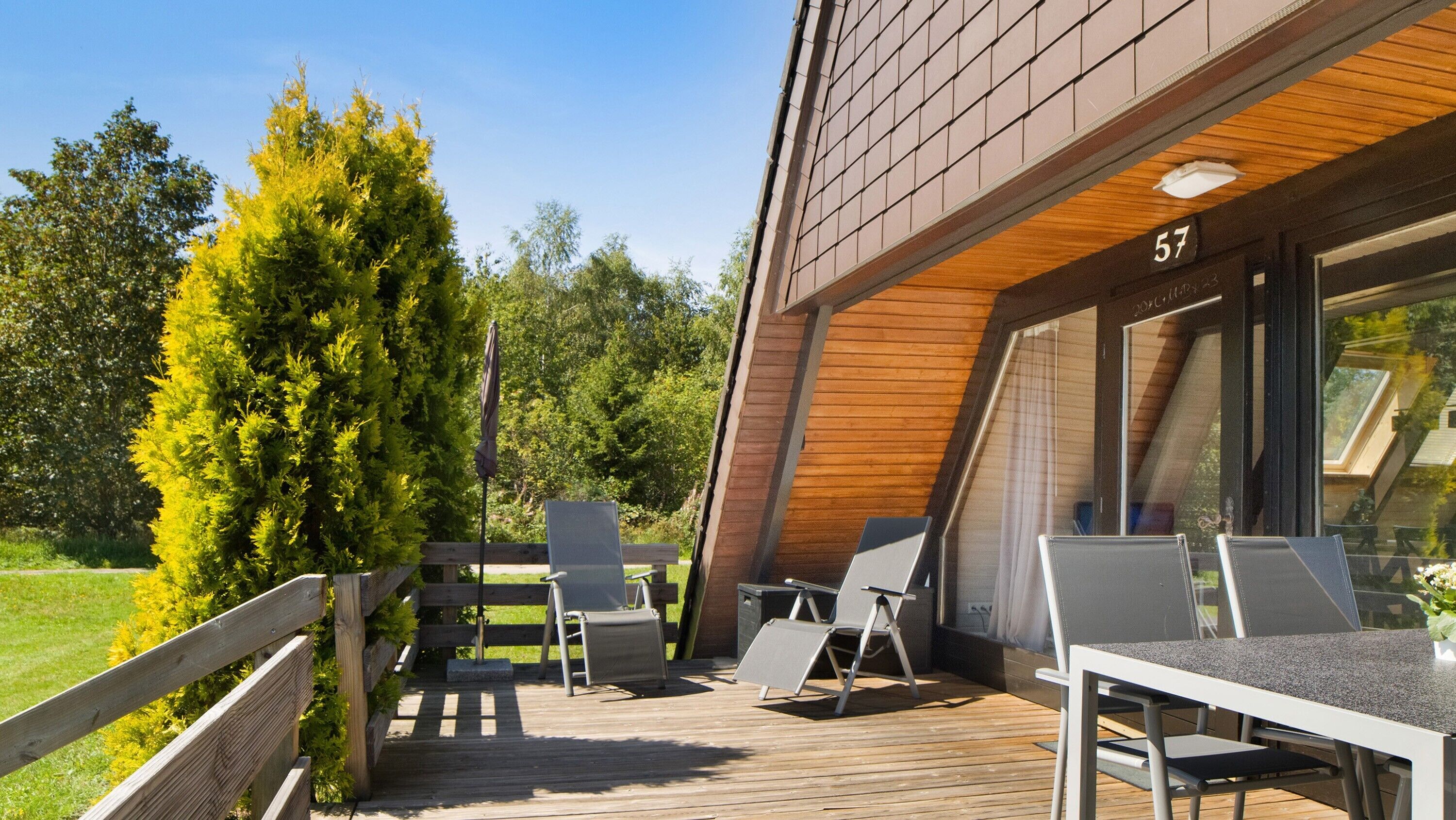 Una terraza de madera con mobiliario exterior, una puerta de cristal de entrada y el número '57' en la pared.