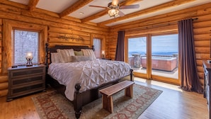 A log cabin bedroom with a large bed, wooden headboard, a nightstand, a bench, and a view of the outside.
