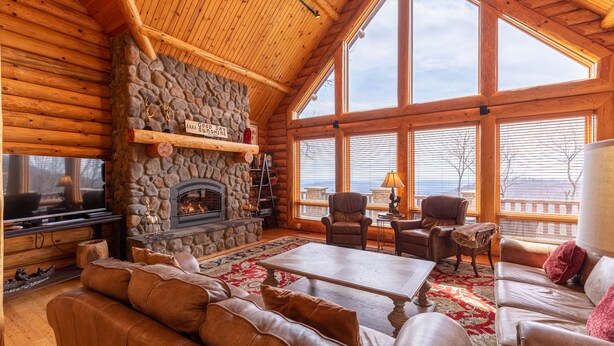 A log cabin interior with a stone fireplace, leather sofas, a television, and large windows offering a view.