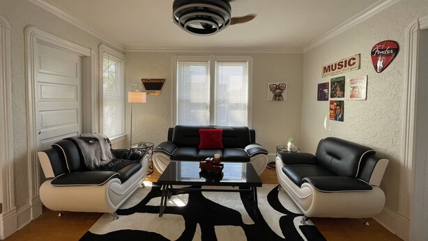 Ein Wohnzimmer mit zwei Sofas, einem Couchtisch und einem Deckenventilator. Zwei Fenster mit Jalousien, eine Wanduhr und ein Schild mit der Aufschrift 'Music'.