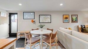 A modern living room with a round dining table, cushioned chairs, a sofa, and a coffee table. There are framed pictures on the wall, a small plant on the table, and a door leading outside.