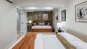 A bedroom with two beds, a desk, and a nightstand. There are framed pictures on the wall and a plant on the nightstand.