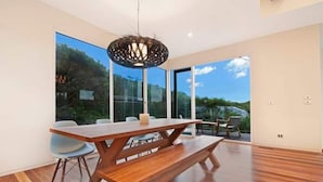 Un espace repas moderne avec une table et des bancs en bois, de grandes fenêtres donnant sur l’extérieur et un luminaire suspendu.
