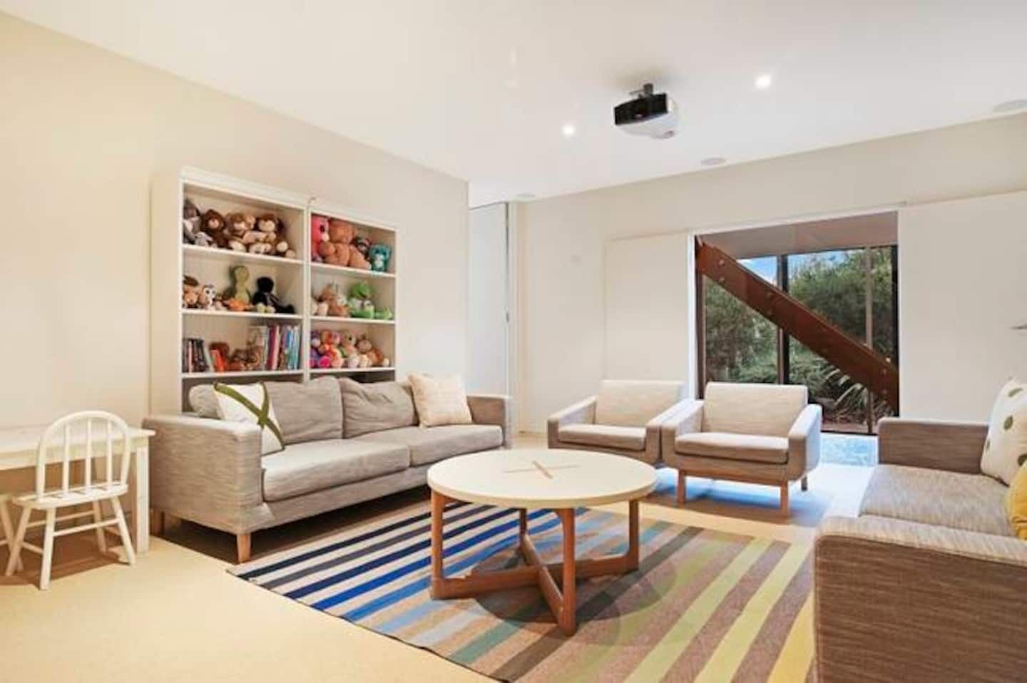 Un salon moderne comprenant un canapé, deux fauteuils, une petite table et une bibliothèque remplie de jouets.