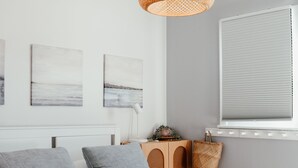 Ein modernes Schlafzimmer mit einem weißen Bett, grauen Kissen, einer türkisblauen Decke, einer gewebten Pendelleuchte und Bildern an der Wand.